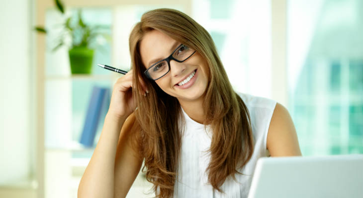 Photo of a woman wearing glasses holding a pen, head tilted to the side, smiling at the camera